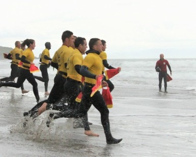 About a dozen first responders wearing wetsuits sprint on a beach toward the ocean water during a water rescue training session.