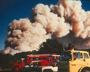 Fire engines parked in the foreground with hills and a massive plume of smoke in the background.