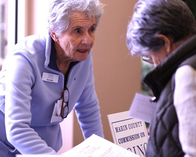 An image of one older woman helping another older woman at a fair booth promoting the Marin County Commission on Aging.