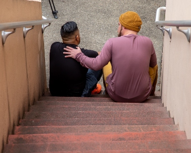 Two men sit on a flight of stairs, with backs facing the camera, and one man's hand is over the shoulder of the other's in a show of support.