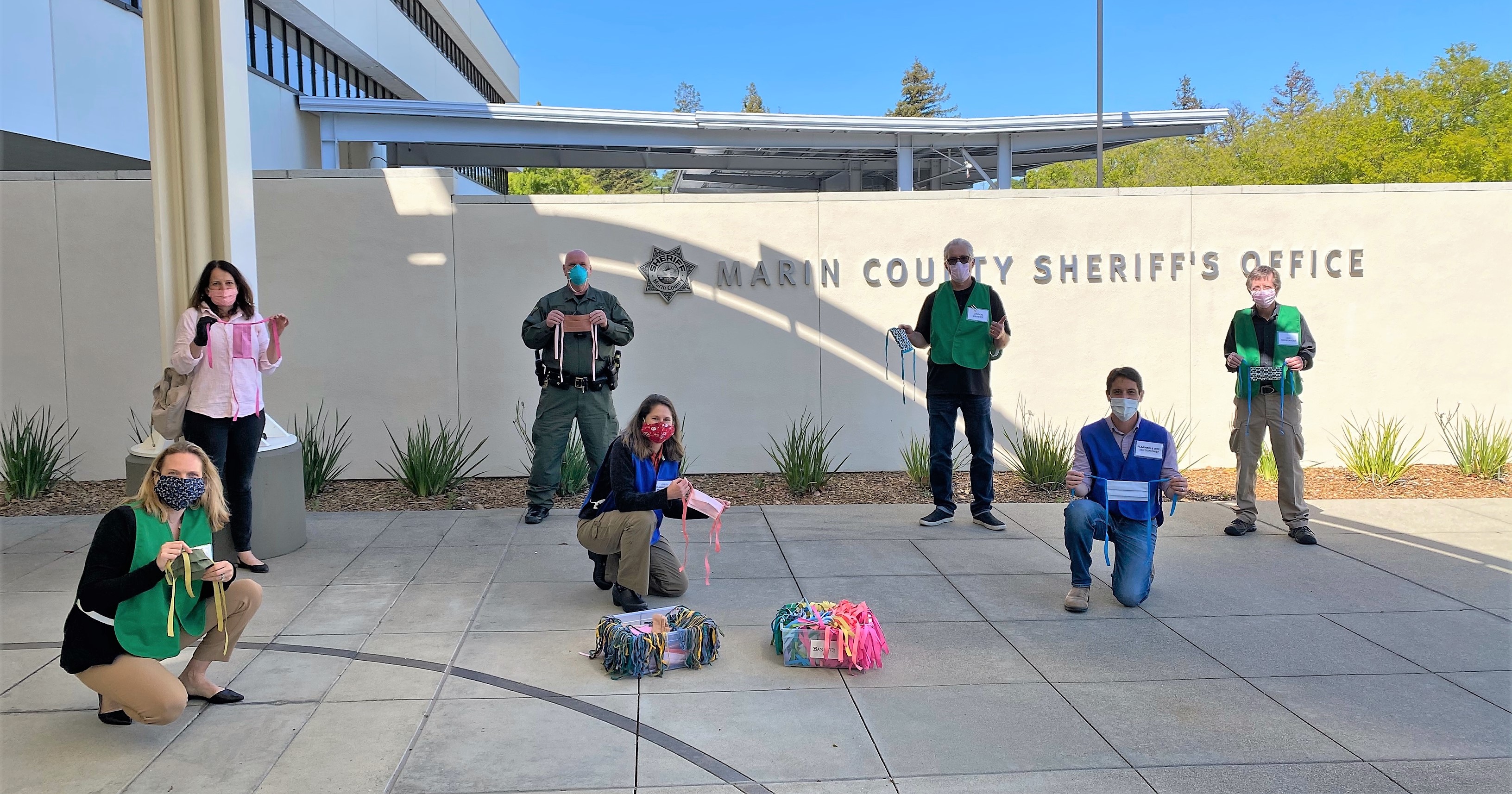 Six County employees wear masks outside the headquarters of the Marin County Sheriff's Office as chief maskmaker Louise Dockstader stands with them.