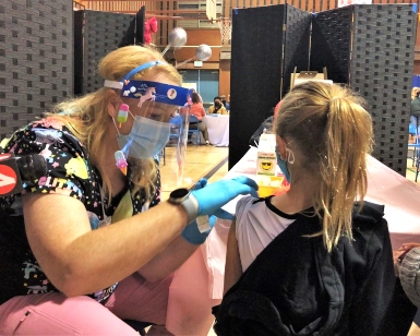 A female nurse on the left leans over to give a COVID-19 vaccine shot to a young girl on the right.