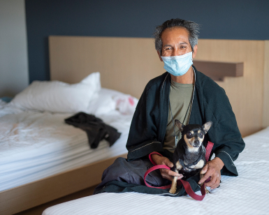 A man in a bathrobe and face covering holds a small dog while sitting on a motel bed.
