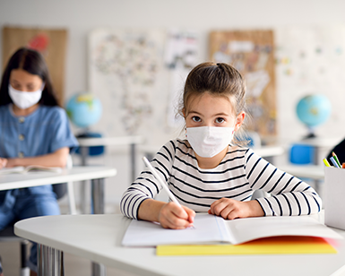 Two female students sit at desks in a classroom, where the desks are spaced more than 6 feet apart and the students are wearing masks.