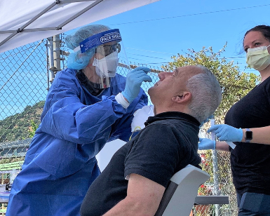 A health care worker in protective gear provides a COVID-19 test to a man sitting in a chair and leaning his head back.