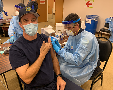 A Marin County Firefighter gives a "thumbs up" while receiving a COVID-19 vaccine from a male nurse.