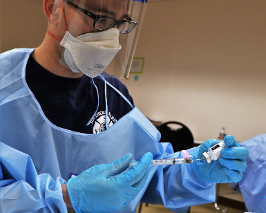 A technician wearing protective gear prepares a dose of the COVID-19 vaccine in a syringe.
