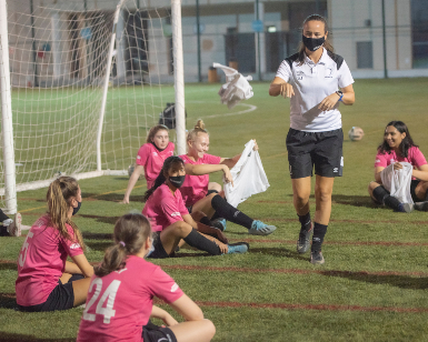 A photo of a female soccer referee wearing a face covering talking to a group of seated female soccer players on the grass.