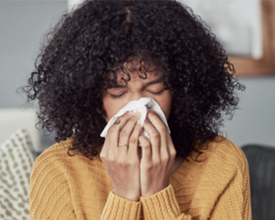 A woman with brown hair and wearing a yellow sweater holds a tissue and blows her nose.  Photo credit: California Department of Public Health.