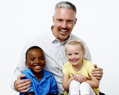 A male preschool teacher sits smiling with a young boy and girl.