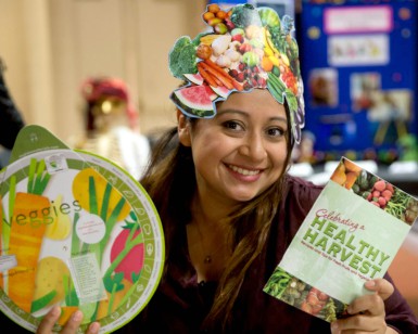 A woman wearing a paper hat with fruit images on it holds up brochures about healthy eating.