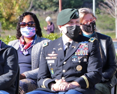 Three people seated at a past military event, including one man wearing full military uniform, all wearing face coverings.