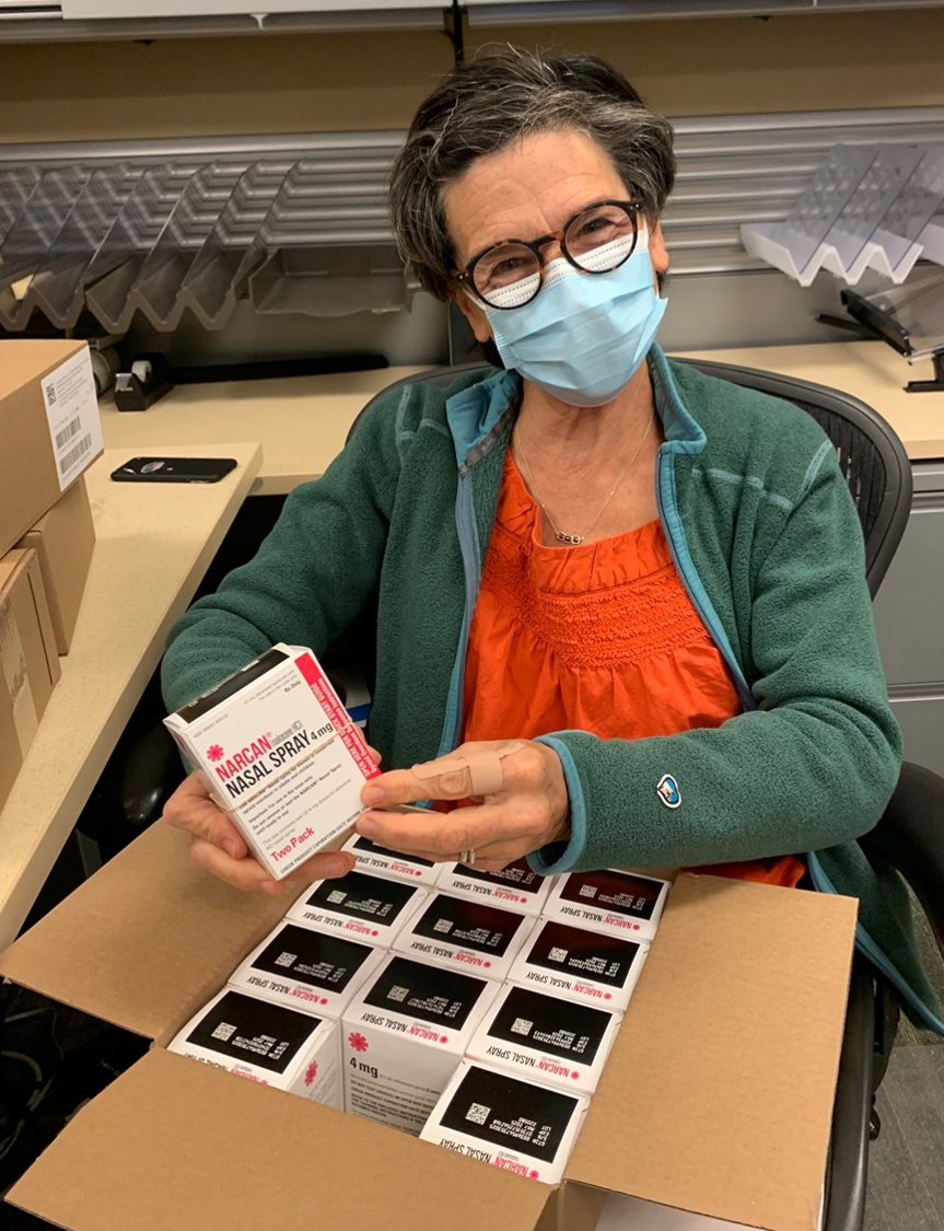 A female volunteer wearing a protective facemask opens a large box of Narcan doses.