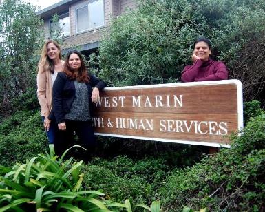 Three women who work at the West Marin Service Center stand next to the large wooden sign outside the building's door.