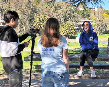 Three teens are showing in a park setting, two on the left videotaping the girl on the right.