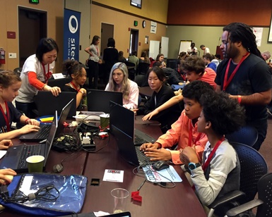 Six teens work on laptop computers as two Hack4Health volunteers assist them.