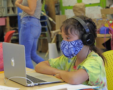A girl of about 9 years old wears a protective mask while looking at a laptop computer 