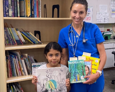 A woman and a young girl hold up books and smile inside a library.