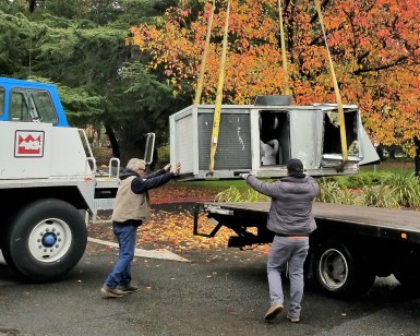 Two workers guide a heavy piece of machinery as it is lifted by a hoist from a truck.