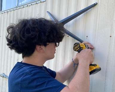 A young person holds a cordless drill and attaches a piece of tech equipment to the outside wall of a building.
