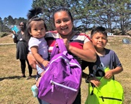 A young mother holds up a summer backpack while holding a very young daughter and standing next to a young son.