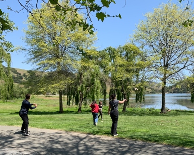 Four teens play in a park with a pond in the background.