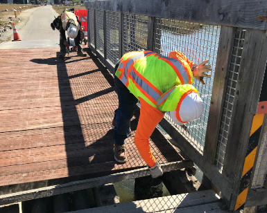 Two workers with construction hats make repairs to a multiuse pathway bridge over a creek.