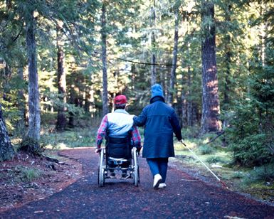 A man pushes himself in a wheelchair and a woman holding a white cane walks beside him on a paved trail in a forest