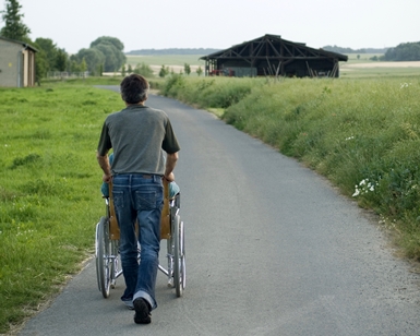A man pushes another person in a wheelchair along a paved pathway in a grassy open space area.