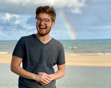 2020 Career Explorer Noah Block is shown smiling on a beach with the ocean and a rainbow in the background. 