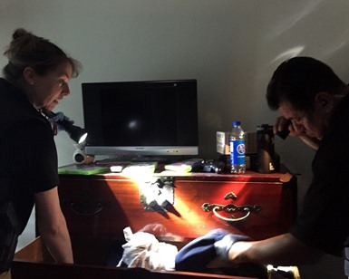 Two law enforcement officers check a sexual offender's chest of drawers in a bedroom.