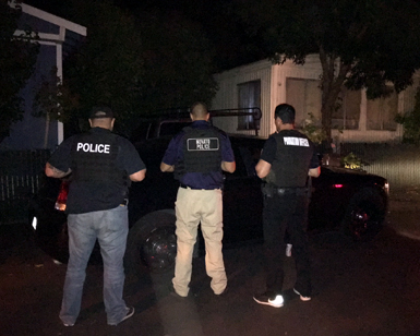 Three law enforcement agencies, with their backs shown, talk to somebody sitting in a car at night.