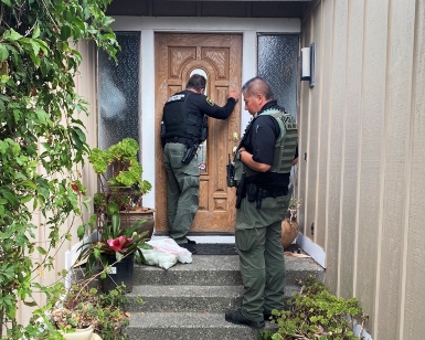 Two male law enforcement officers knock on a front door of the home of a past DUI offender. 