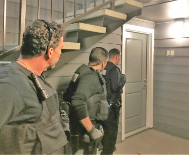 Three law enforcement officers in full protective gear get ready to knock on a front door.