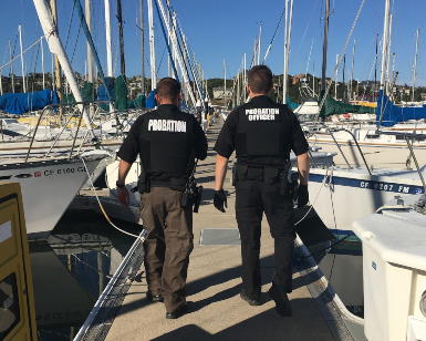 Two male probation officers walk away from the camera on a dock with boats on either side of them.