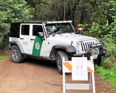 A Sheriff's Office Jeep is parked to block an unpaved trail with signage in front of the vehicle about maintaining social distancing during the COVID-19 pandemic.